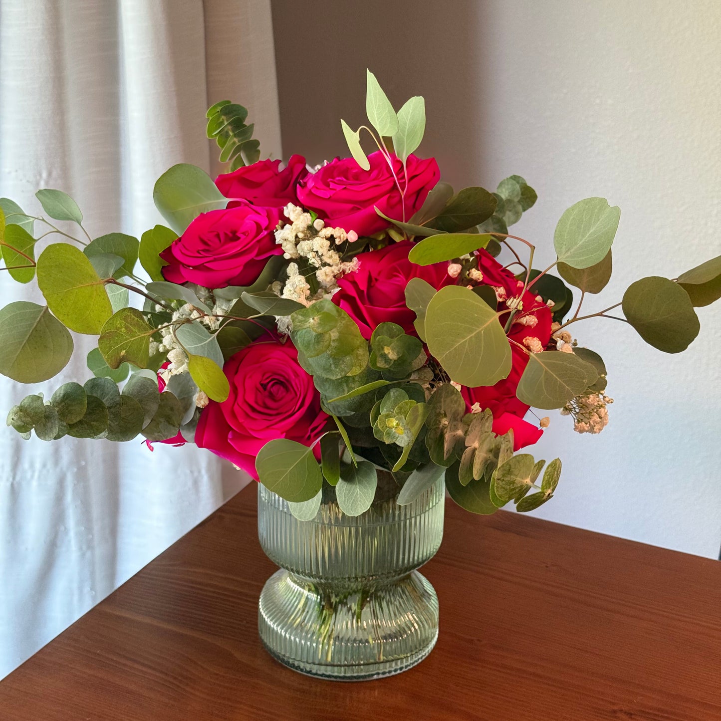 Bouquet of redpinkroses and greenery in a clear vase on a wooden surface with a white curtain background.