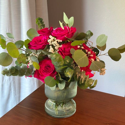 Bouquet of redpinkroses and greenery in a clear vase on a wooden surface with a white curtain background.