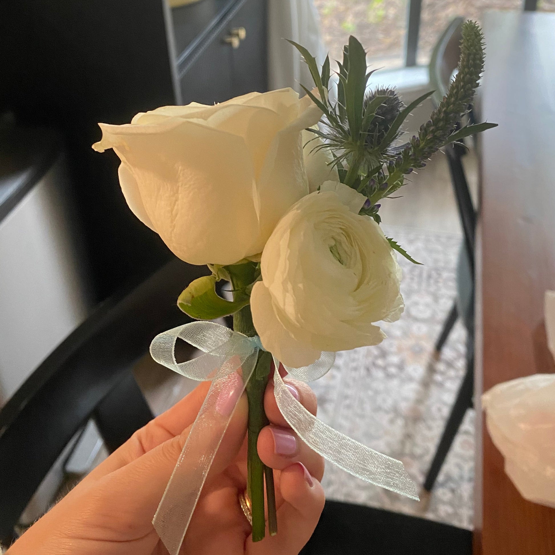 White rose boutonniere with green leaves held by a hand in front of a window.